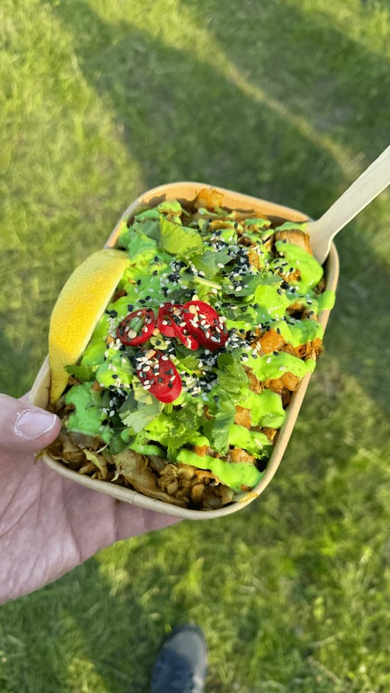 A small carton of food with a fork in it. The carton contains chicken and is covered in a green sauce with chillis and seeds sprinkled on top. A lemon slice is placed at the side.